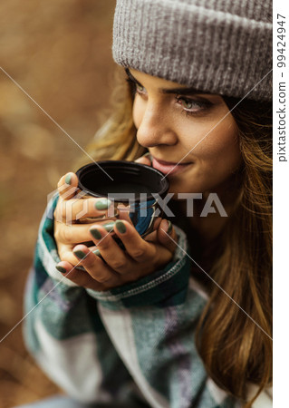 Smiling millennial caucasian woman in jacket and hat drink hot tea cup, travel in cold forest, enjoy adventure outdoor Smiling millennial caucasian woman in jacket and hat drink hot tea cup, travel in cold forest, enjoy adventure outdoor 99424947