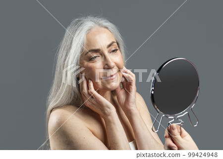 Cheerful old european lady with gray hair touches face with fingers, looks in mirror, enjoy result Cheerful old european lady with gray hair touches face with fingers, looks in mirror, enjoy result 99424948