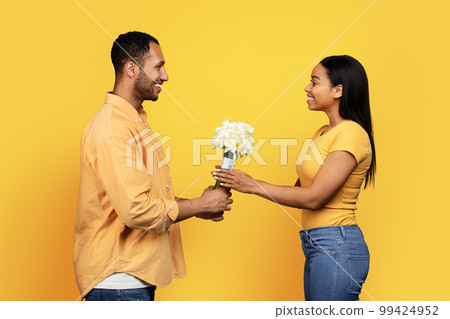 Smiling young african american guy gives bouquet of flowers to woman, isolated on yellow background, studio Smiling young african american guy gives bouquet of flowers to woman, isolated on yellow background, studio 99424952