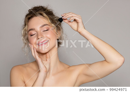 Young beautiful lady applying facial serum, using anti-aging treatment product, posing over grey studio background Young beautiful lady applying facial serum, using anti-aging treatment product, posing over grey studio background 99424971