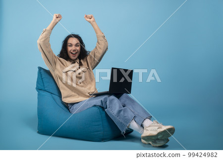 Laughing shocked excited millennial european female student sitting on bag chair with laptop, raising hands up 99424982