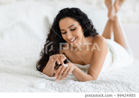 Happy millennial caucasian brunette lady with long hair lies on soft bed, applies cream on hands Happy millennial caucasian brunette lady with long hair lies on soft bed, applies cream on hands 99424985