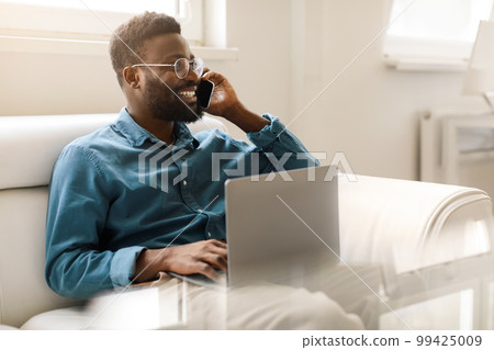 Happy black businessman in glasses calling by phone, working with laptop on couch in office interior, free space 99425009