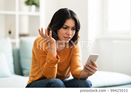 Frustrated arab woman looking at smartphone while sitting on couch at home 99425015