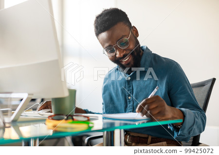 African american male entrepreneur working on modern computer and taking notes, sitting at workpace in office interior African american male entrepreneur working on modern computer and taking notes, sitting at workpace in office interior 99425072