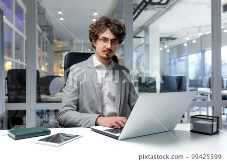 Portrait of thinking serious businessman inside office, bearded man looking into camera focused in glasses, worker in business clothes sitting at workplace using laptop at work. 99425599