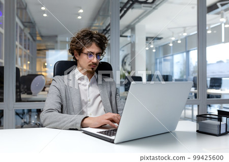 Serious and focused businessman inside modern office working on laptop, bearded man typing on keyboard, thinking man in business clothes sitting at workplace. 99425600