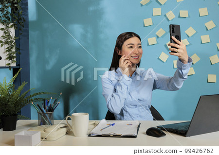 Corporate asian female employee sitting at contemporary office desk taking selfie with smart phone. Professional woman smiling in front of mobile device screen at work station. 99426762