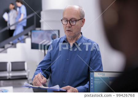 Elderly man holding clipboard signing medical documents before start consultation with doctor in hospital waiting area. Receptionist is managing patient records and scheduling follow-up appointments. 99426768
