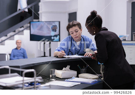 African american reception worker checking medical report with assistant discussing patient expertise while working at medication treatment during checkup visit in hospital lobby. Medicine concept 99426826