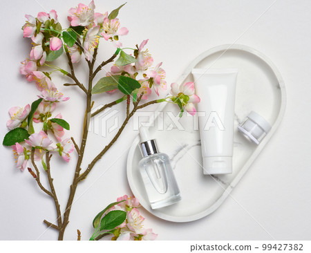 White plastic tub, glass bottle with a dropper on a marble stand, and a bouquet of blooming cherry on a white background, cosmetic container, top view 99427382