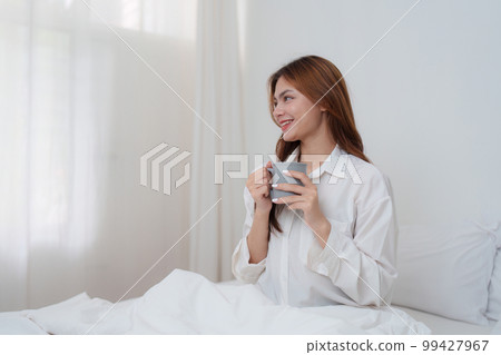 Beautiful asian woman has woken up on a white bed and holding coffee in the morning at home. lifestyle concept. 99427967