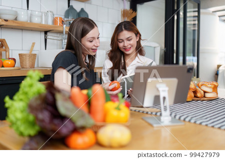 Beautiful woman and friend talking and using laptop at kitchen room. Girl friends couple living together at home. 99427979