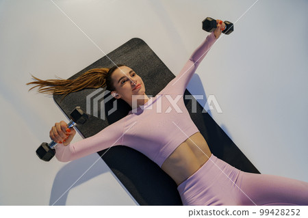 Top view of strong woman lying on yoga mat and doing exercises for hands muscles with dumbbells 99428252