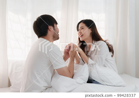 Beautiful asian couple in love and smiling sitting on bed. Romantic moment, relationships, family concept. 99428337