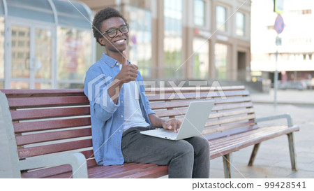 Young African Man with Laptop Showing Thumbs Up Sign While Sitting Outdoor on Bench Young African Man with Laptop Showing Thumbs Up Sign While Sitting Outdoor on Bench 99428541