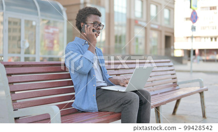 Young African Man Talking on Phone and using Laptop while Sitting Outdoor on Bench Young African Man Talking on Phone and using Laptop while Sitting Outdoor on Bench 99428542