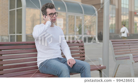 Young Man Talking on Phone while Sitting Outdoor on Bench Young Man Talking on Phone while Sitting Outdoor on Bench 99428601