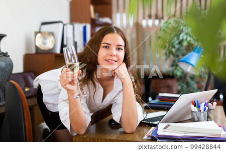 Cheerful young female with glass of champagne posing at office Cheerful young female with glass of champagne posing at office 99428844