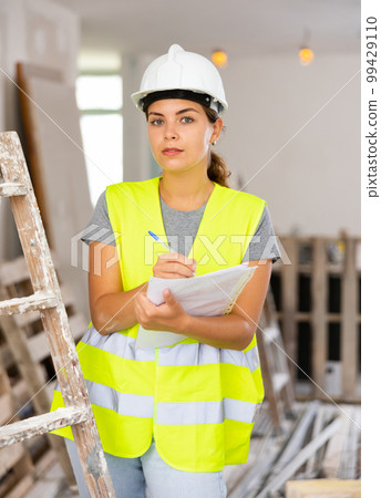 Woman foreman in a protective helmet and yellow vest checks the completed construction work on drawing 99429110