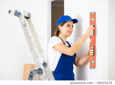 Satisfied young woman builder checking vertical of walls with level Satisfied young woman builder checking vertical of walls with level 99429236