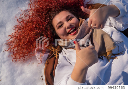 Top view of a fat red-haired woman lying on the snow. 99430563