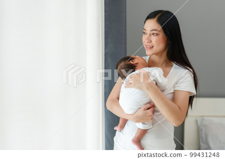 mother holding and consoling newborn baby to sleeping near the window in bedroom 99431248