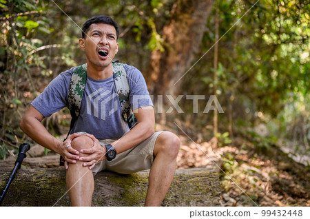 A stressed Asian male trekker sits on a wooden log and holds his knee, severe leg pain, injured A stressed Asian male trekker sits on a wooden log and holds his knee, severe leg pain, injured 99432448