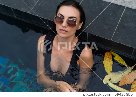Beautiful european woman in black swimsuit posing outside at villa in swimming pool, on vacation, perfect shape, good body, tanned, fit, wearing trendy brown sunglasses 99433890