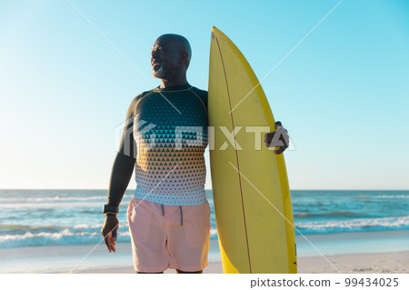 African american senior man with surfboard looking away while standing against sea and clear sky 99434025