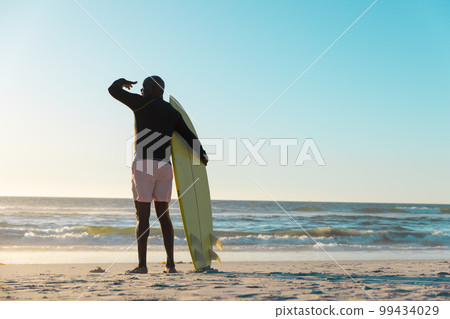 Rear view of african american senior man with surfboard standing on beach and looking at sea and sky Rear view of african american senior man with surfboard standing on beach and looking at sea and sky 99434029