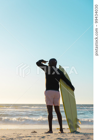 Rear view of senior african american man with surfboard looking at sea under clear sky at sunset Rear view of senior african american man with surfboard looking at sea under clear sky at sunset 99434030
