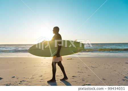 Side view of african american senior man with surfboard walking on sandy beach against sea and sky Side view of african american senior man with surfboard walking on sandy beach against sea and sky 99434031