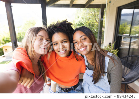 Portrait of happy diverse teenage female friends embracing on balcony taking selfie 99434245