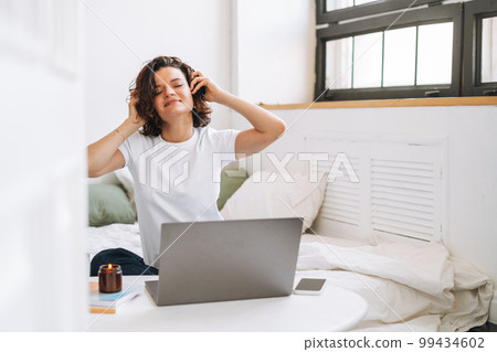 Young brunette woman with closed eyes in headphones working on laptop from home 99434602