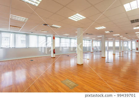 Large empty space with ceiling tiles, fluorescent lights, light brown laminate flooring and white painted columns. Large office space left from a company. 99439701