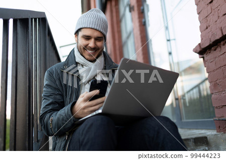 a young man sits on the steps with a laptop and looks at the phone with a smile a young man sits on the steps with a laptop and looks at the phone with a smile 99444223