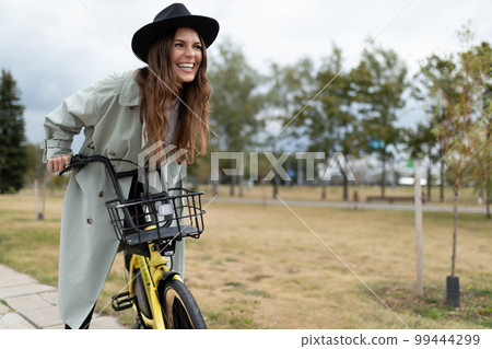 cheerful young woman in a gray coat and black hat rides a bike with a smile cheerful young woman in a gray coat and black hat rides a bike with a smile 99444299