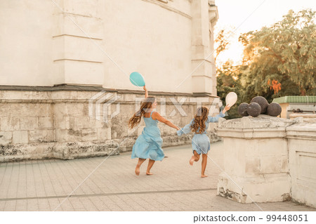 Daughter mother run holding hands. In blue dresses with flowing long hair, they hold balloons in their hands against the backdrop of a sunset and a white building. 99448051