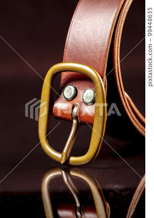 Part of a brown leather belt with a buckle on a dark background. 99448635