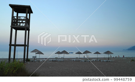 An evening view of a sandy beach with umbrellas, sunbeds and a lifeguard tower. People swimming in the sea, walking along the beach. The Mediterranean coast. An evening view of a sandy beach with umbrellas, sunbeds and a lifeguard tower. People swimming in the sea, walking along the beach. The Mediterranean coast. 99448759