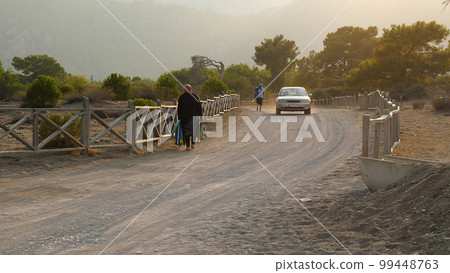 The car is driving along a sandy road surrounded by a wooden fence. People are walking on the beach. In the background, a green forest in the setting sun. The car is driving along a sandy road surrounded by a wooden fence. People are walking on the beach. In the background, a green forest in the setting sun. 99448763