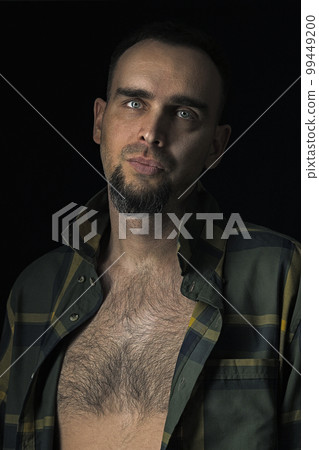 Handsome male model in an unbuttoned checked shirt exposing his hairy torso. Portrait of a bearded Caucasian man against a black background. Sexy man looking into the camera. Handsome male model in an unbuttoned checked shirt exposing his hairy torso. Portrait of a bearded Caucasian man against a black background. Sexy man looking into the camera. 99449200