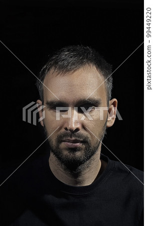 Portrait of a middle-aged man against a black background. A serious man with grey hair and a small beard stares thoughtfully to the side. A Caucasian man wearing a black T-shirt. Portrait of a middle-aged man against a black background. A serious man with grey hair and a small beard stares thoughtfully to the side. A Caucasian man wearing a black T-shirt. 99449220