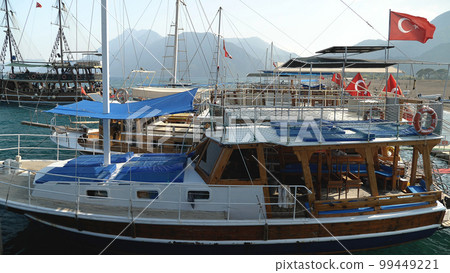 View from a boat of a wooden pleasure craft anchored off the coast. Turkish flags are hoisted on it. In the background are mountains and blue sea. A summer holiday in the Mediterranean. View from a boat of a wooden pleasure craft anchored off the coast. Turkish flags are hoisted on it. In the background are mountains and blue sea. A summer holiday in the Mediterranean. 99449221