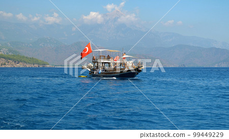 View from the stern of a wooden pleasure boat with tourists. In the background high, beautiful mountains, clouds and blue sea. The Turkish flag is flying on the boat. View from the stern of a wooden pleasure boat with tourists. In the background high, beautiful mountains, clouds and blue sea. The Turkish flag is flying on the boat. 99449229