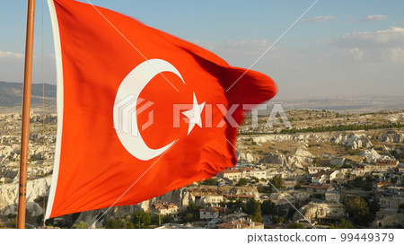 The Turkish flag in Cappadocia. The Turkish flag waving in the wind against a background of mountains and houses. Close-up. The Turkish flag in Cappadocia. The Turkish flag waving in the wind against a background of mountains and houses. Close-up. 99449379