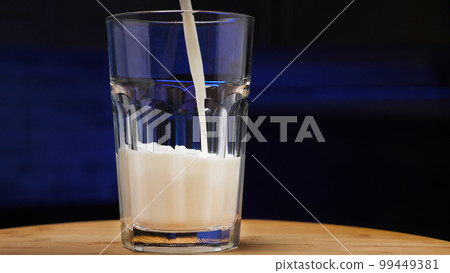 Pouring milk into a glass. Glass of milk on a wooden tray. Dark blue background. The concept of milk products. Pouring milk into a glass. Glass of milk on a wooden tray. Dark blue background. The concept of milk products. 99449381