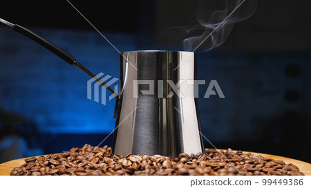 A pile of roasted brown coffee beans and a steel cezve. Steam rises from the jezve. Turkish coffee. Coffee shop concept. A pile of roasted brown coffee beans and a steel cezve. Steam rises from the jezve. Turkish coffee. Coffee shop concept. 99449386
