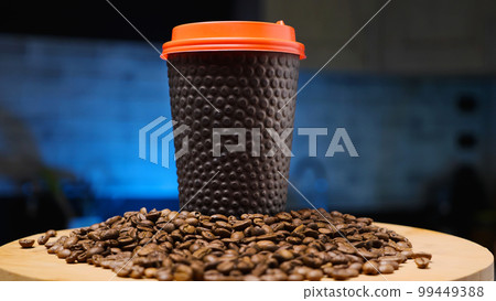 A black paper coffee cup with a red lid surrounded by roasted coffee beans. Dark blue background. Coffee shop concept. 99449388